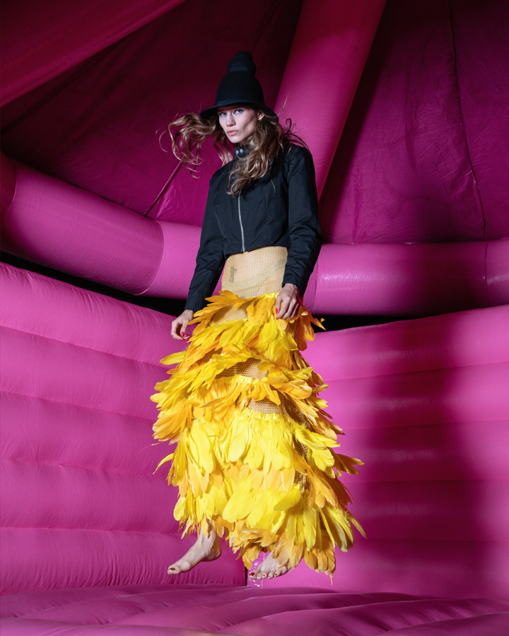 Model in bright yellow dress and black jacket posing mid-air in a bright pink bounce house